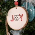 A round wooden Christmas ornament with the word "joy" painted in red letters