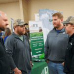 Four men speak to each other while conference attendees explore informational booths nearby