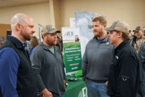 Four men speak to each other while conference attendees explore informational booths nearby