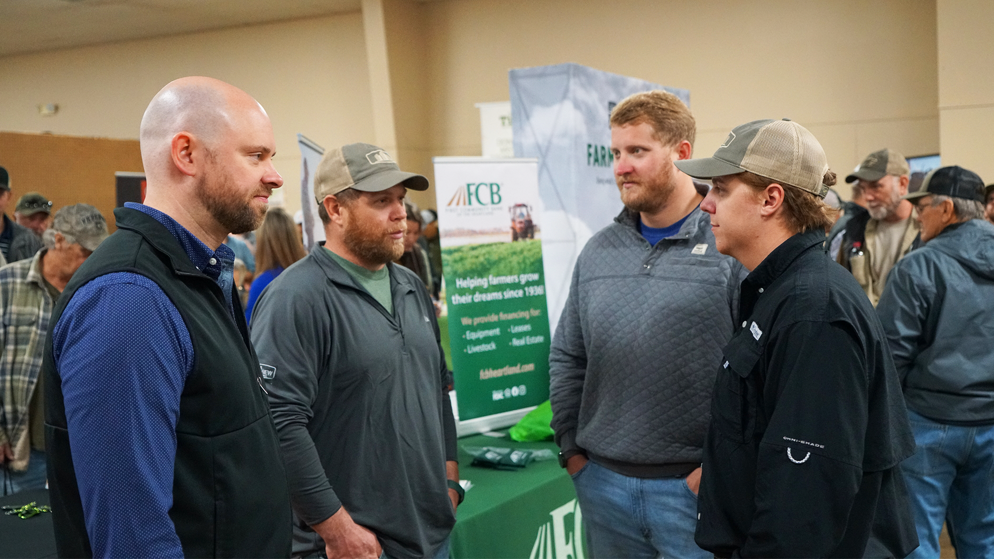 Four men speak to each other while conference attendees explore informational booths nearby