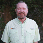 A man in a green shirt smiles for a photo in front of a row of trees