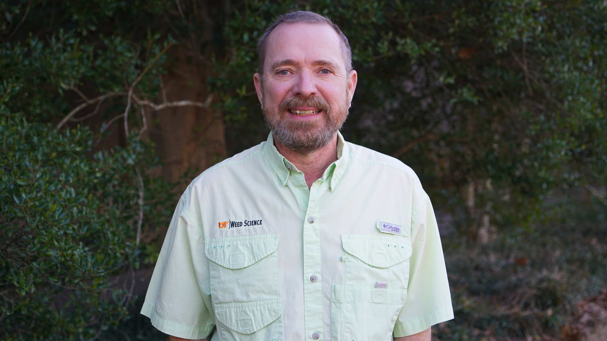 A man in a green shirt smiles for a photo in front of a row of trees