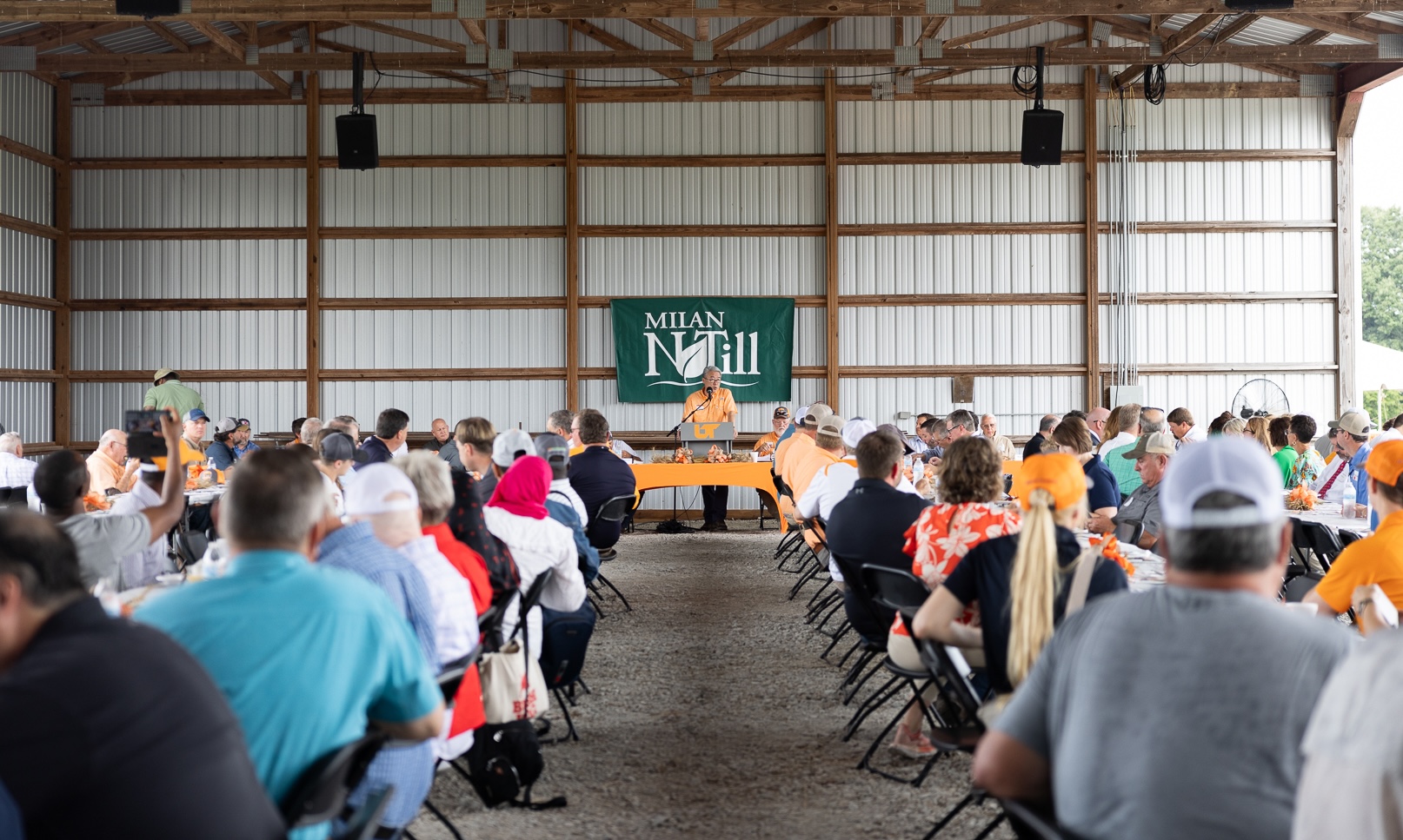 Tables of attendees listen to a speaker at a podium with a "Milan No-Till" banner them