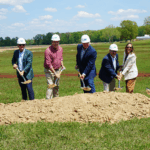 A line of officials wearing formal attire and white hard hats pose for a photo while shoveling dirt from a small pile