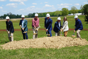 A line of officials wearing formal attire and white hard hats pose for a photo while shoveling dirt from a small pile