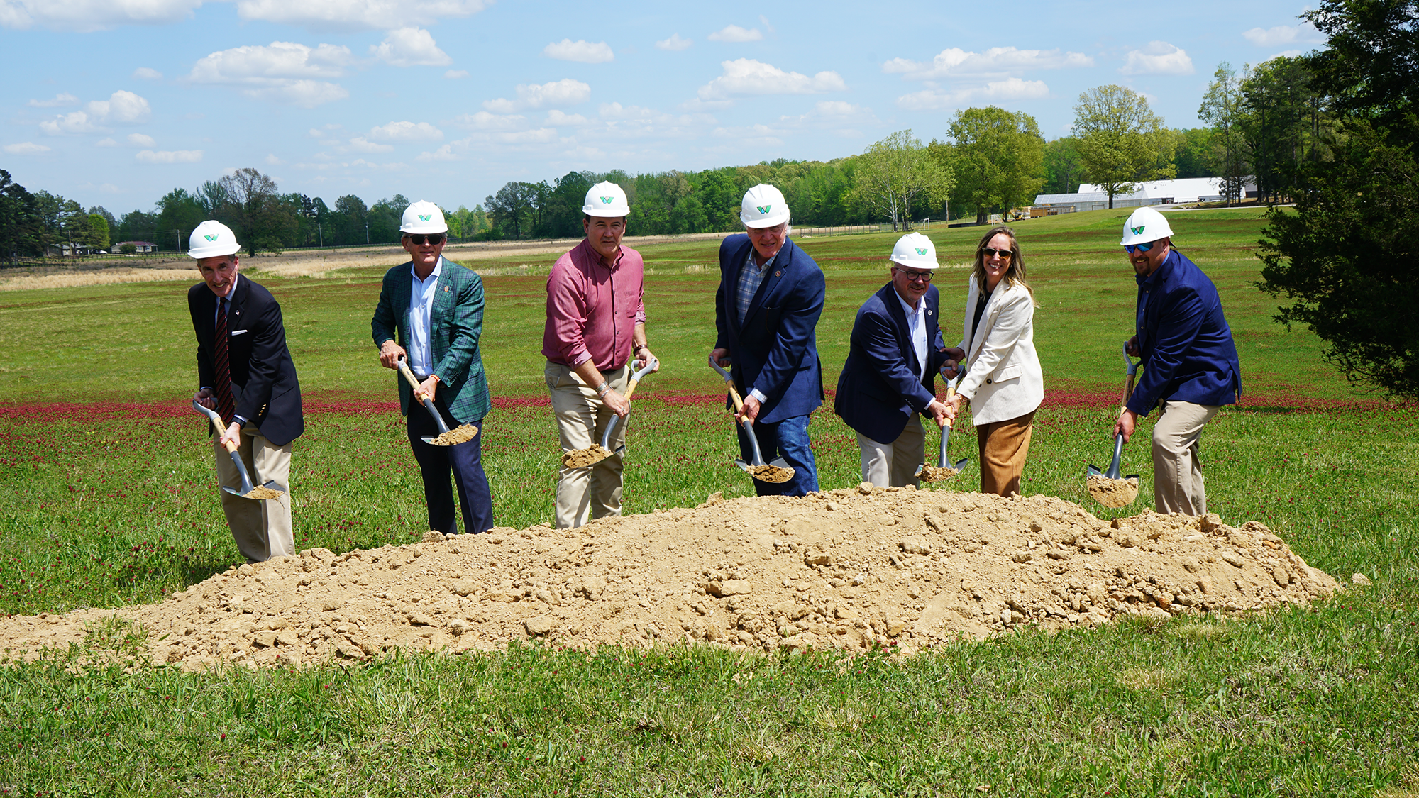A line of officials wearing formal attire and white hard hats pose for a photo while shoveling dirt from a small pile