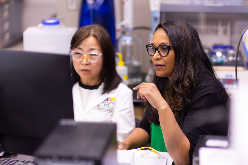 Two individuals in a laboratory setting are focused on a computer screen, with one gesturing while they discuss data or results.