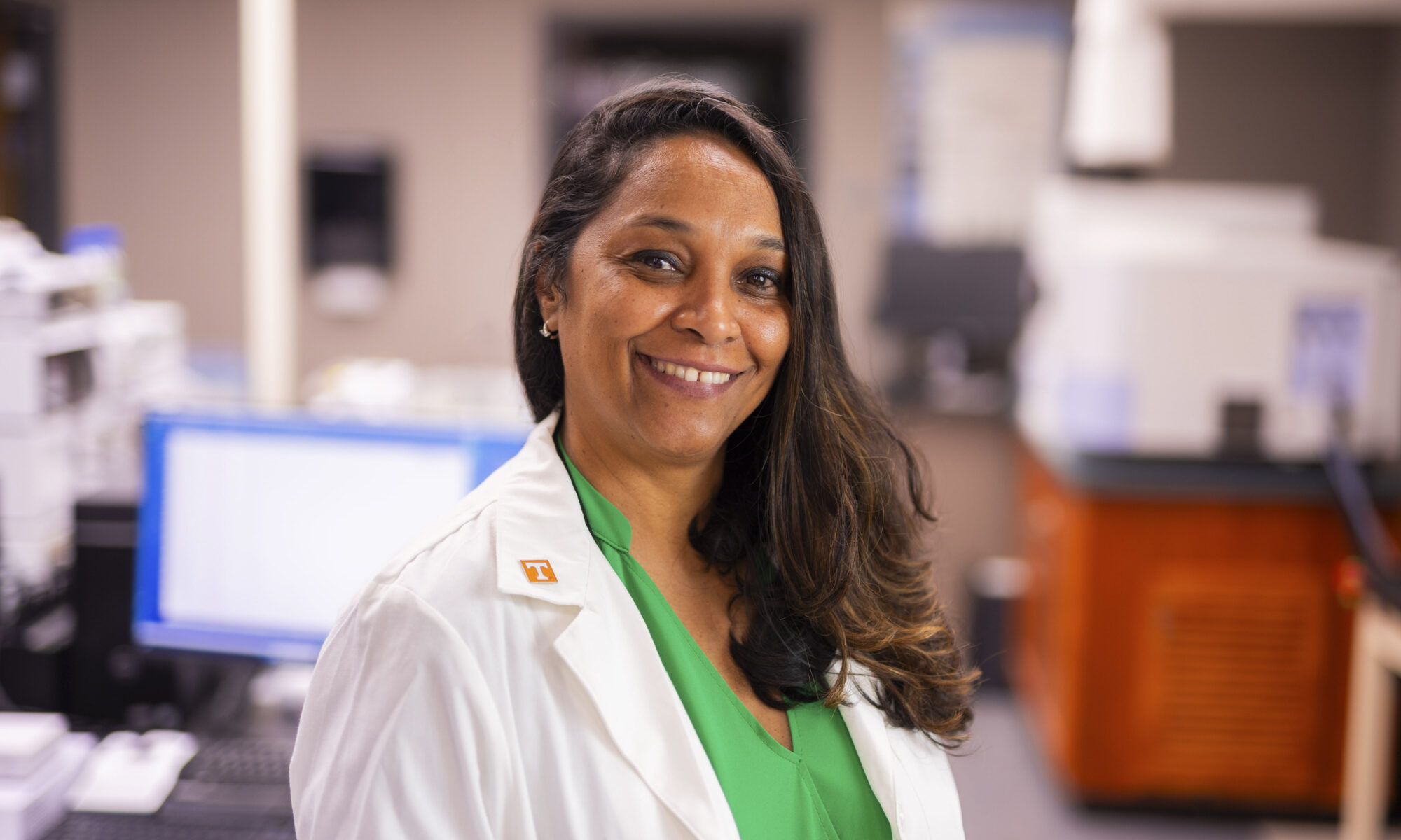 A smiling individual in a lab coat stands in a laboratory setting, wearing a green shirt and a white coat with a "T" pin, surrounded by scientific equipment.