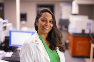 A smiling individual in a lab coat stands in a laboratory setting, wearing a green shirt and a white coat with a