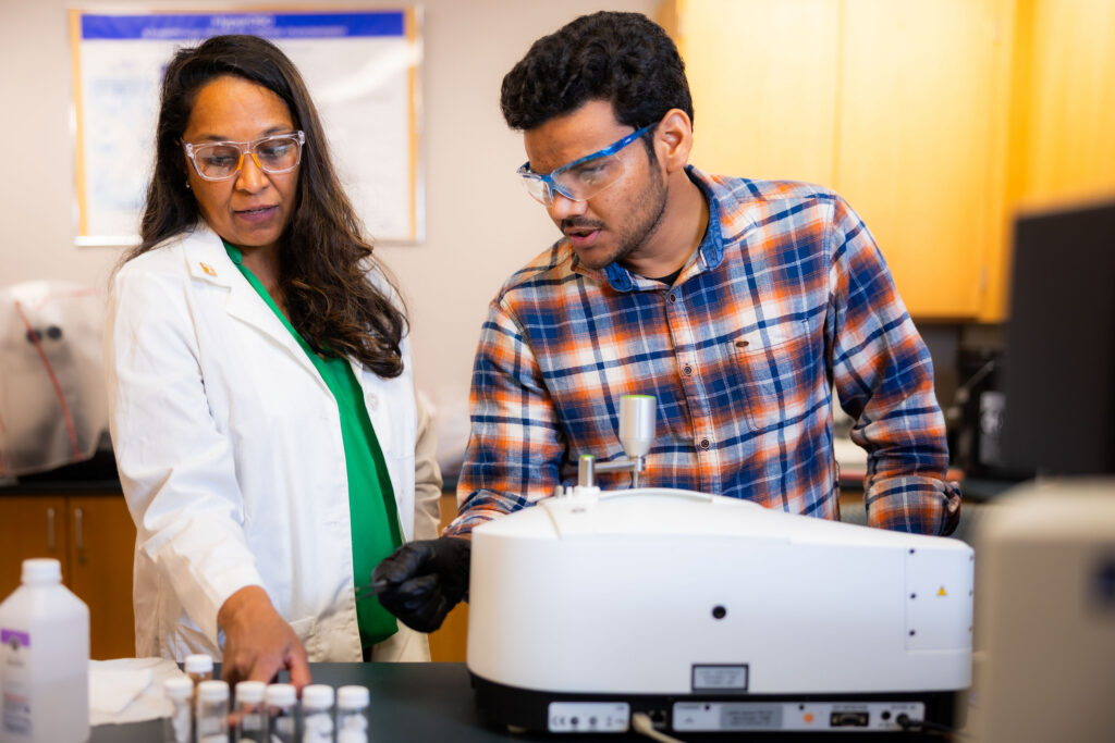 Two individuals in a lab setting, wearing safety goggles, are focused on operating scientific equipment.