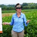 A woman in a cap and blue shirt presents poster boards in front of a soybean field