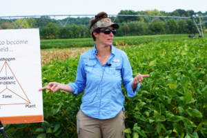 A woman in a cap and blue shirt presents poster boards in front of a soybean field