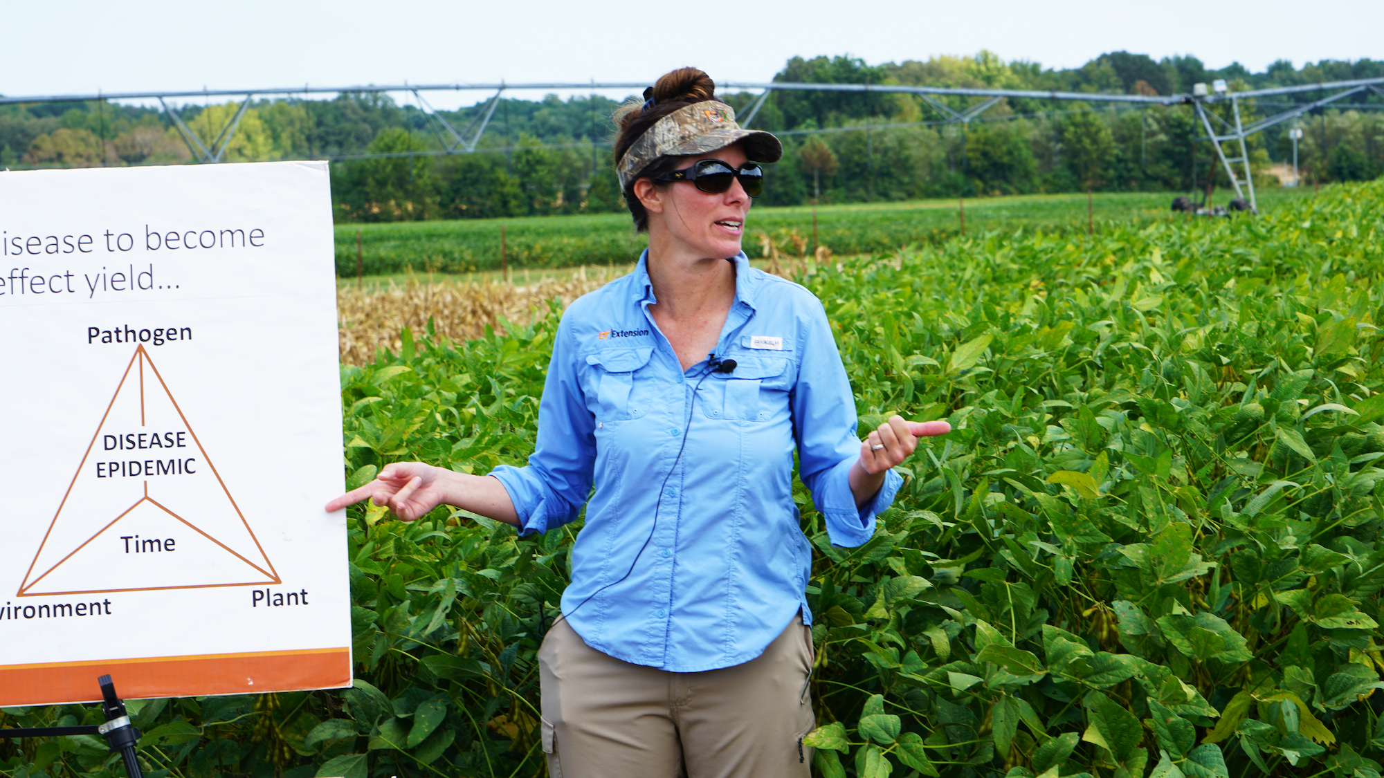 A woman in a cap and blue shirt presents poster boards in front of a soybean field