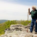 A man holding a walking stick stands on the side of a cliff overlooking the horizon.