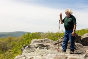A man holding a walking stick stands on the side of a cliff overlooking the horizon.