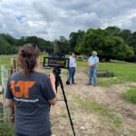 A woman films two men talking in a field.