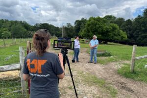 A woman films two men talking in a field.