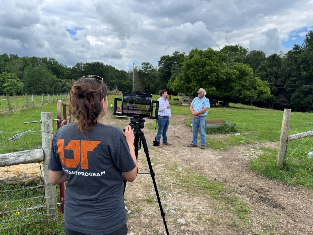 A woman films two men talking in a field.
