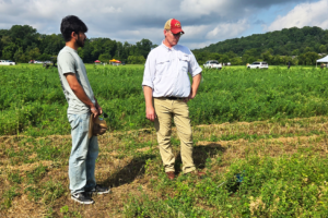 A student holding a clipboard stands next to an instructor in a hat and white shirt while observing a weed research plot.