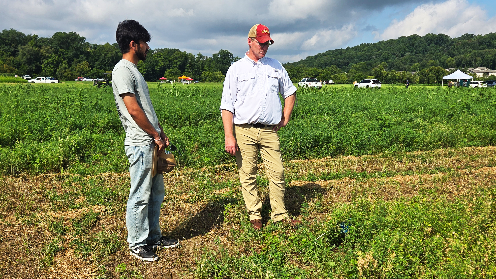 A student holding a clipboard stands next to an instructor in a hat and white shirt while observing a weed research plot.