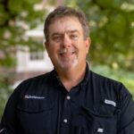 A man in a black button shirt smiles while posing for a photo
