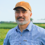 A man in a hat and blue shirt poses for a headshot in front of a crop field
