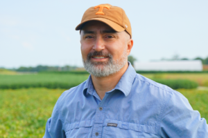 A man in a hat and blue shirt poses for a headshot in front of a crop field