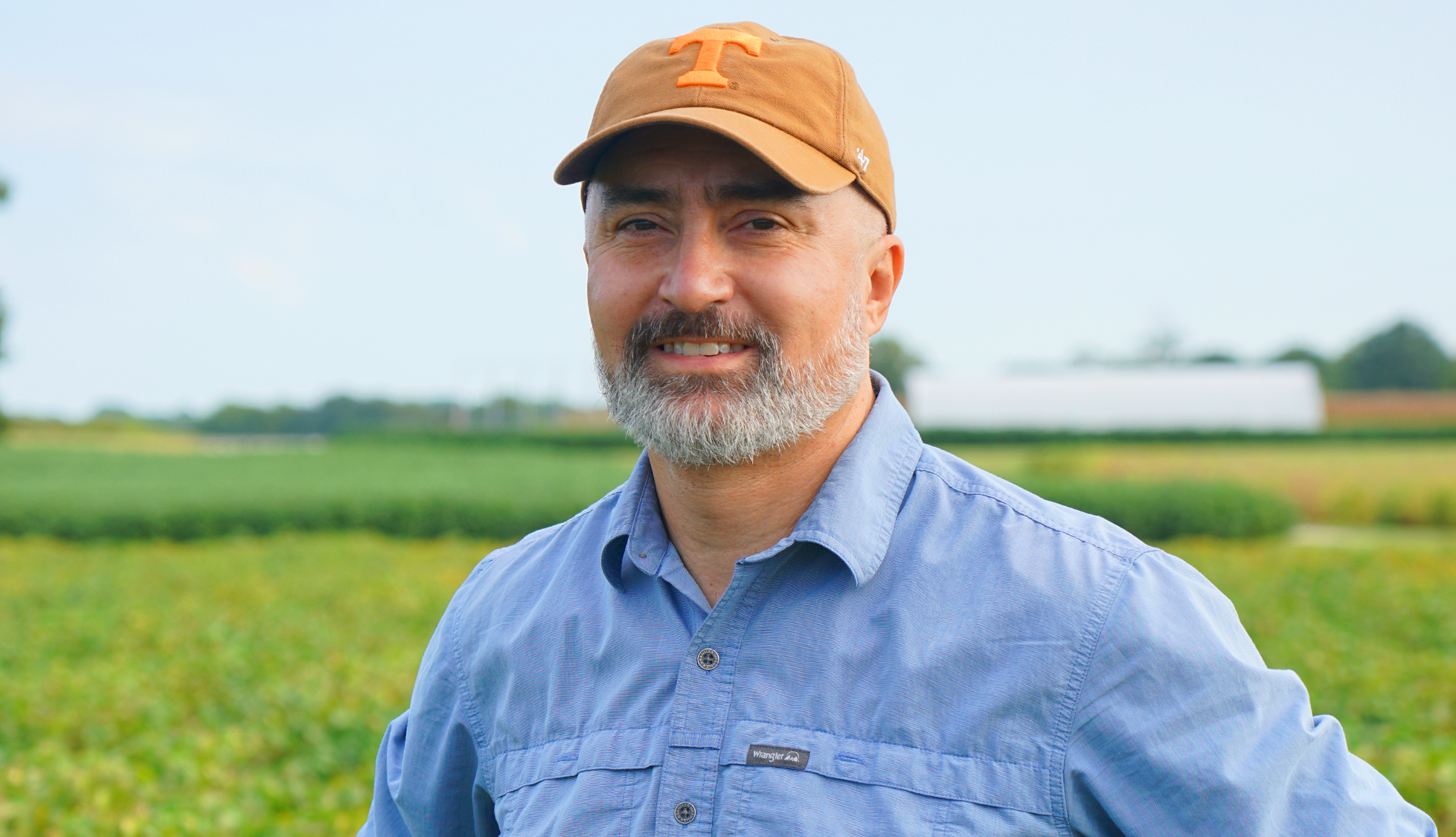 A man in a hat and blue shirt poses for a headshot in front of a crop field